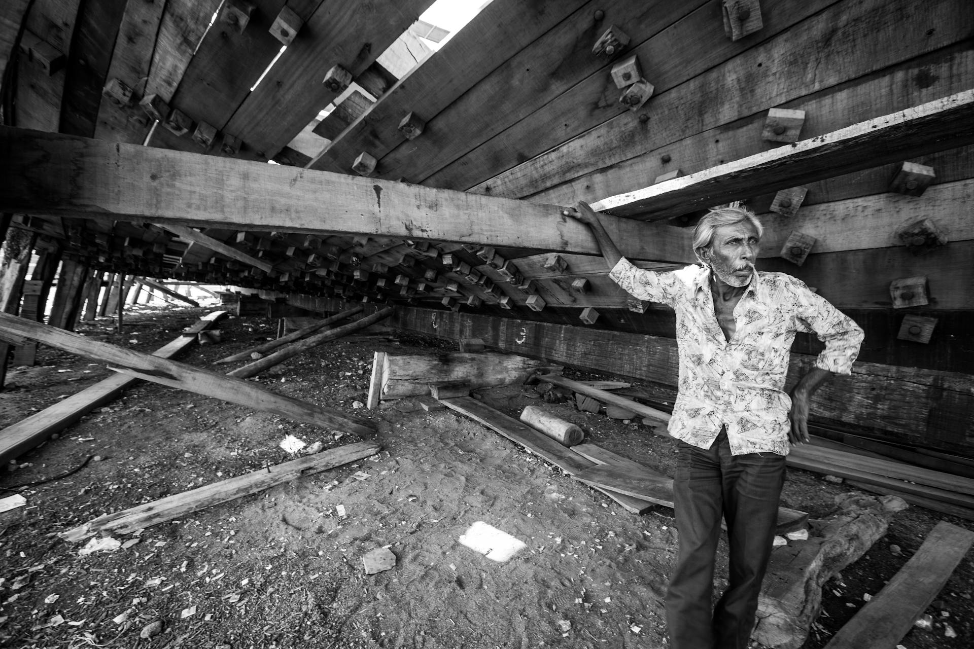 An elderly man leans against a wooden ship structure in Mandvi shipyard, India, showcasing traditional craftsmanship.