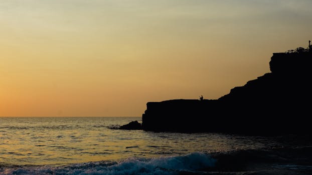 Silhouette of a person fishing on Bali's cliffside as the sun sets over the ocean.
