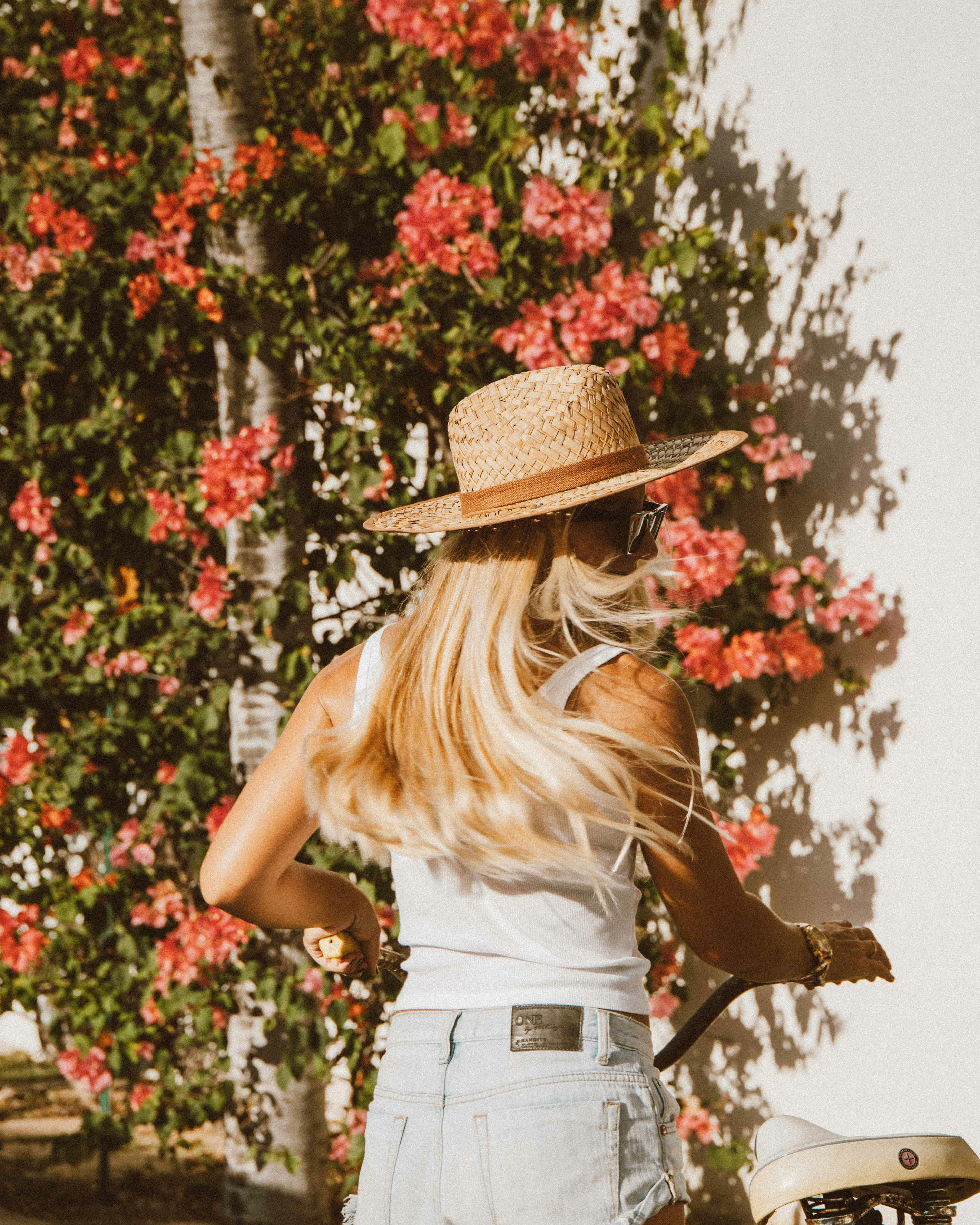 在一个阳光明媚的日子里，一位留着金色长发、头戴草帽的女子走过盛开的花丛，展现出充满活力与时尚的生活场景