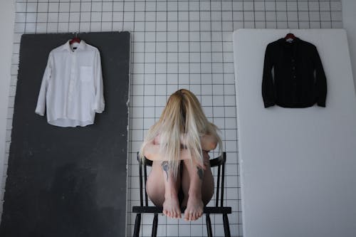 Unrecognizable barefoot depressed tattooed female embracing knees while sitting on stool behind black and white shirts on hangers