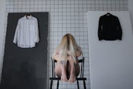 一位赤脚、纹身、神情沮丧的女性坐在凳子上，她身后挂着一件黑白衬衫，正抱着膝盖