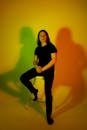 Man with long hair in total black look siting on chair and illuminated by multicolored light against background of colored shadows