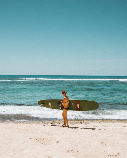 Free Woman in Black Bikini Holding White Surfboard Walking on Beach Stock Photo