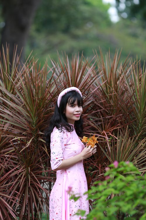 Free Side view of young content Asian lady in stylish apparel and head rim standing with flowers near plant and looking at camera Stock Photo