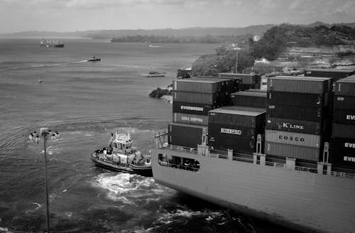 Black and white contemporary cargo vessel with many containers floating on calm river near hilly grassy shore towards industrial port