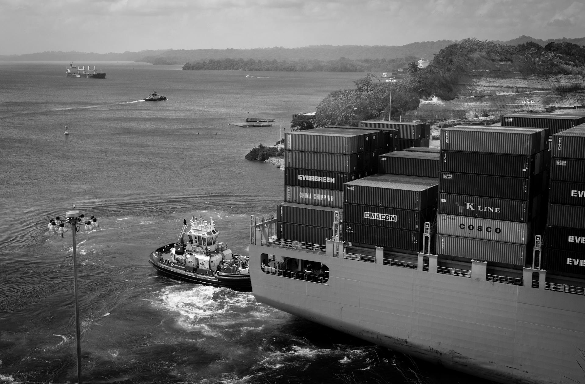 Black and white contemporary cargo vessel with many containers floating on calm river near hilly grassy shore towards industrial port
