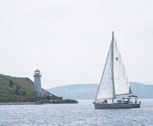 Scenery of modern small sailboat floating on calm rippled seawater near green hilly coastline with white lighthouse on summer day