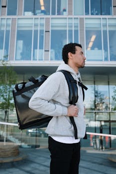 Young male courier with backpack in front of a modern city building, ready for delivery.