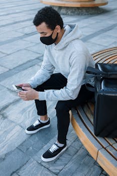 A young man in a hoodie and mask sitting on a bench using a smartphone, with a delivery bag.