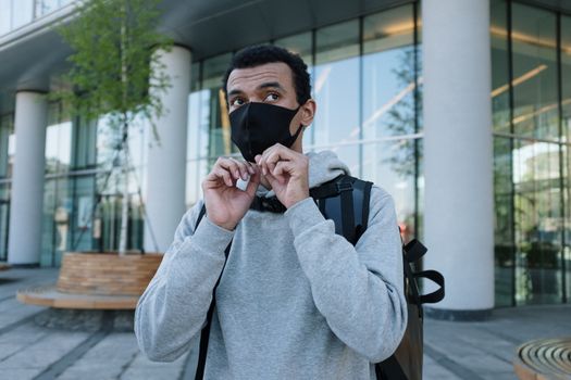 Black male courier adjusts mask in modern urban environment for safe delivery.