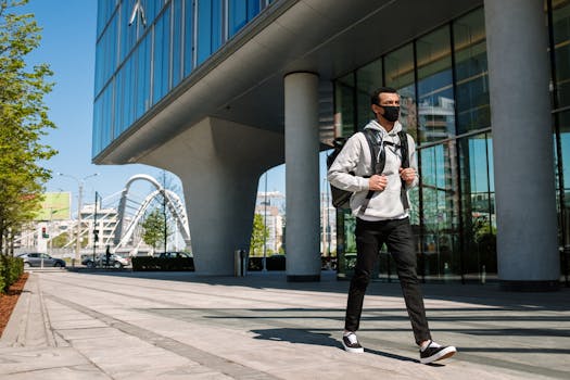 Courier with backpack and face mask walks by sleek urban architecture in daytime.