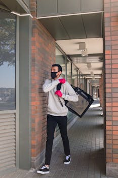Courier in a face mask and gloves delivers food outdoors, walking by brick buildings.
