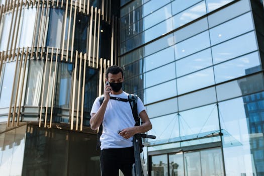 Man with black mask and backpack stands with an electric scooter in a modern city setting.