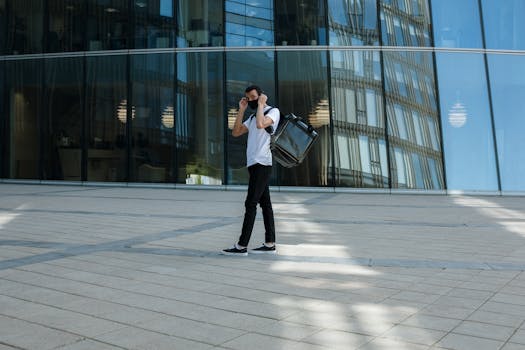 A young adult mall courier wearing a face mask carries a delivery backpack outside a sleek glass office building.
