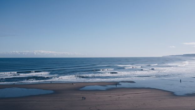 Amazing scenery of people walking on sandy coastline washed by peaceful calm seawater under cloudless blue sky on summer day
