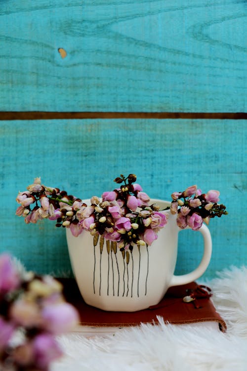 Free Arrangement of white mug with tender tiny pink flowers placed on diary on white fluffy coverlet near wooden blue wall Stock Photo