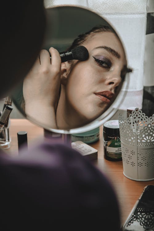 Crop woman looking in mirror and applying blush on face