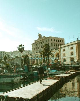 Back view faceless male in casual wear walking along wooden dock across old tropical city canal on sunny weather