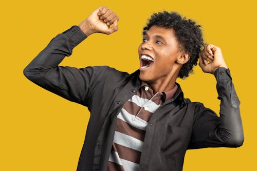 Expressive African American male wearing casual clothes standing against yellow background in studio and raising fists in triumph while looking away and screaming happily
