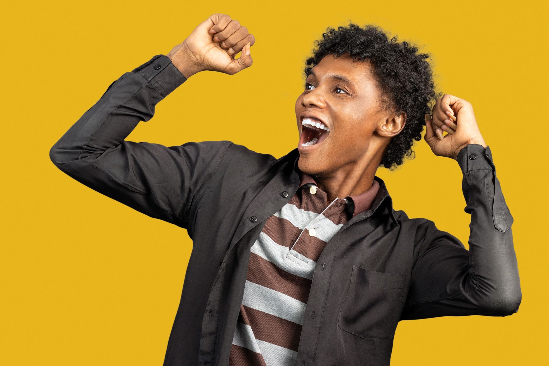 Expressive African American male wearing casual clothes standing against yellow background in studio and raising fists in triumph while looking away and screaming happily