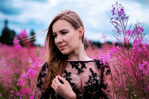 Charming young female in black lace dress standing amidst bright pink flowers blossoming in lush field and looking away dreamily