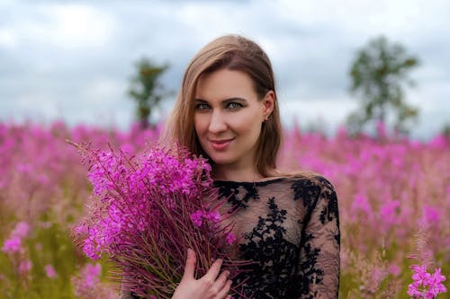 Glad young female wearing elegant black dress standing with delicate pink flowers in hands on blooming field and looking at camera contentedly