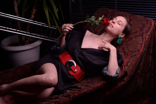 一位性感的年轻女性闭着眼睛坐在深红色椅子上，身穿时髦的黑色连衣裙，用红玫瑰轻抚嘴唇