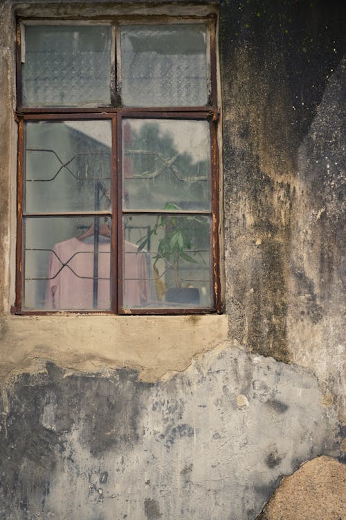 Through glass of various clothes hanging on rack in shabby stone house with dirty weathered walls and old wooden window