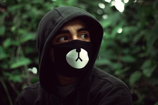 Portrait of a man in a bear face mask and black hoodie surrounded by green foliage.