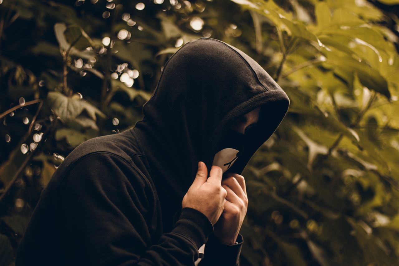a person wearing a hoodie and face mask stands in a natural setting ca