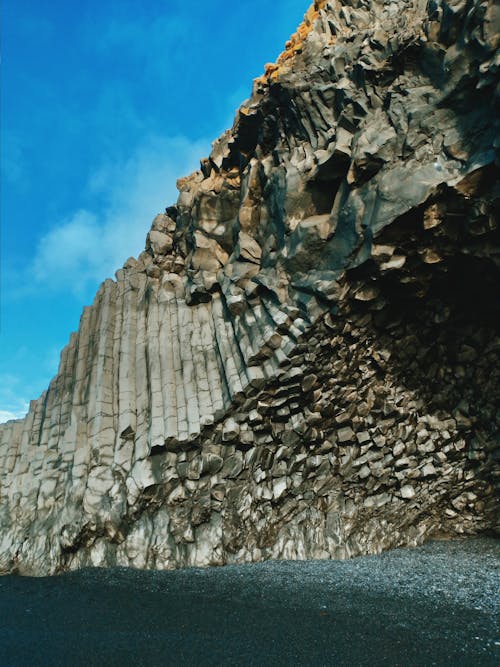 Vibrant blue sky with clouds above dark massive unusual cliff above wet pebbles