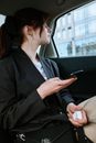 一个扎着马尾辫、戴着耳环的女人正坐在汽车后座，专注地用智能手机拍着窗外