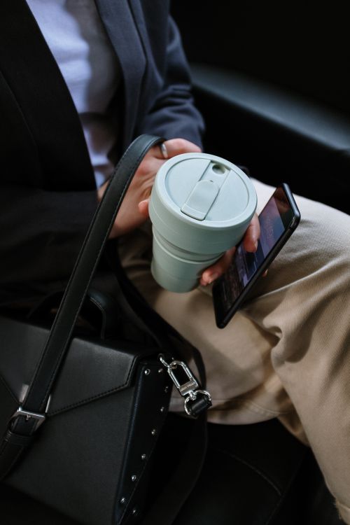 Free Person holding reusable coffee cup and smartphone in a car, depicting eco-friendly commute. Stock Photo