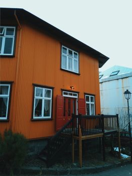 Grey cloudless sky above small brown two storey house with wooden outside stairs between bush and lantern near white construction