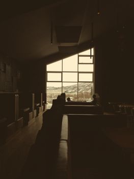 Dark brown tables and couches under angled ceiling in empty dark cafe with mountain through big window