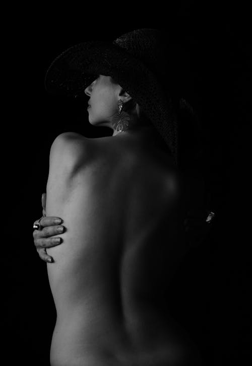 Black and white of young faceless topless female in hat on head touching back and wearing stylish big earring