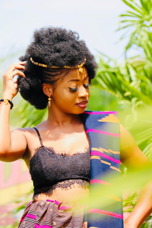 Pretty slim African American curly female with closed eyes in colourful  traditional wear posing sensually and touching hair with hand near green plants
