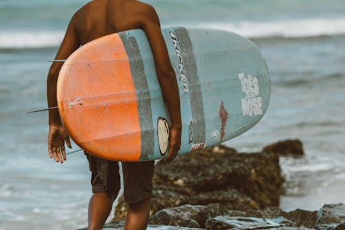 Man Carrying Orange and Blue Surfboard Standing on Rocky Shore