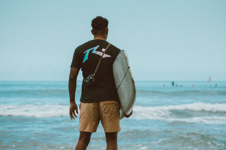A surfer stands with a surfboard on a beach in Honolulu, Hawaii, gazing at the ocean waves.