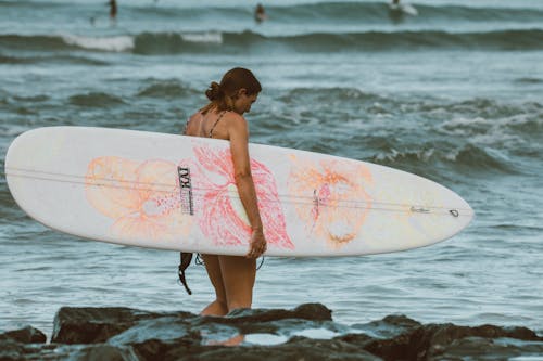 Woman in Pink and White Floral Bikini Holding White Surfboard Standing on Beach