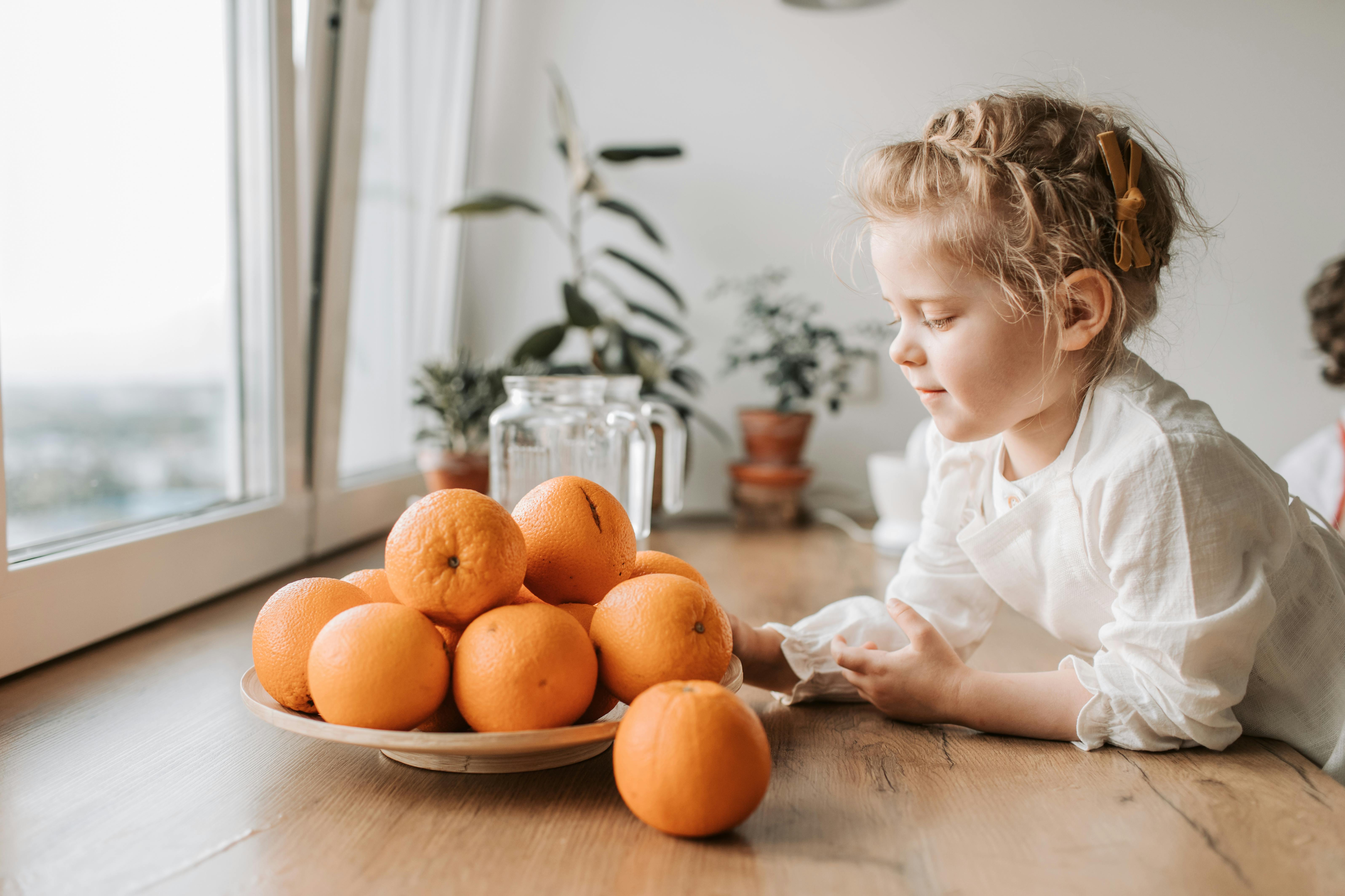 Girl in White Dress Shirt Sitting Beside Orange Fruit on Brown Wooden Table