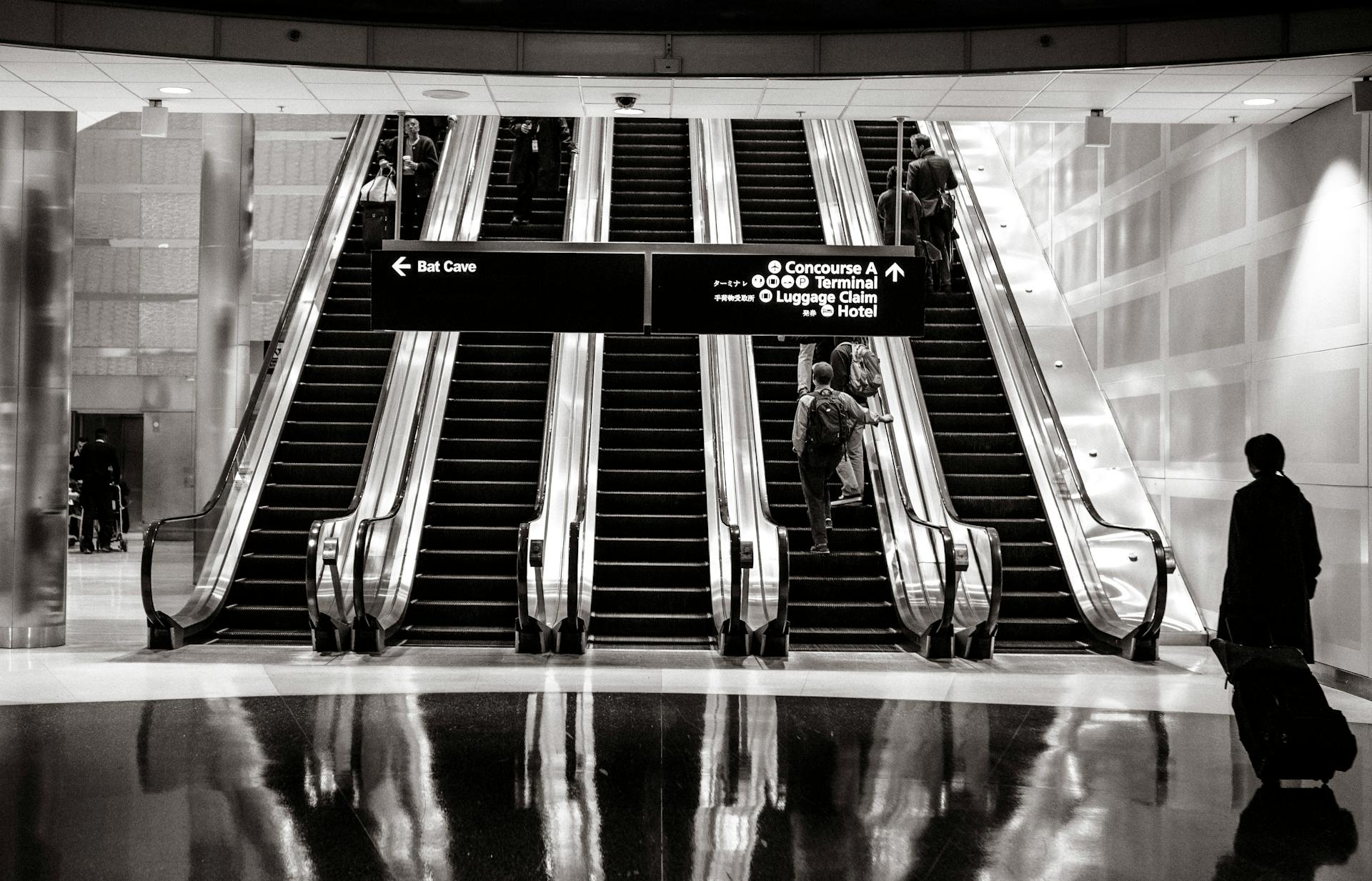 Travelers on escalators in a modern airport terminal with directional signs overhead.