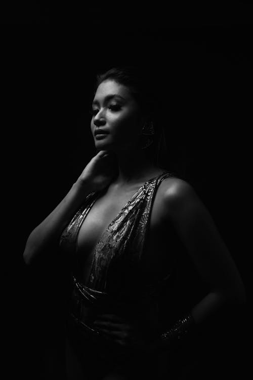 Black and white of young sensual ethnic woman in elegant wear touching neck in darkness