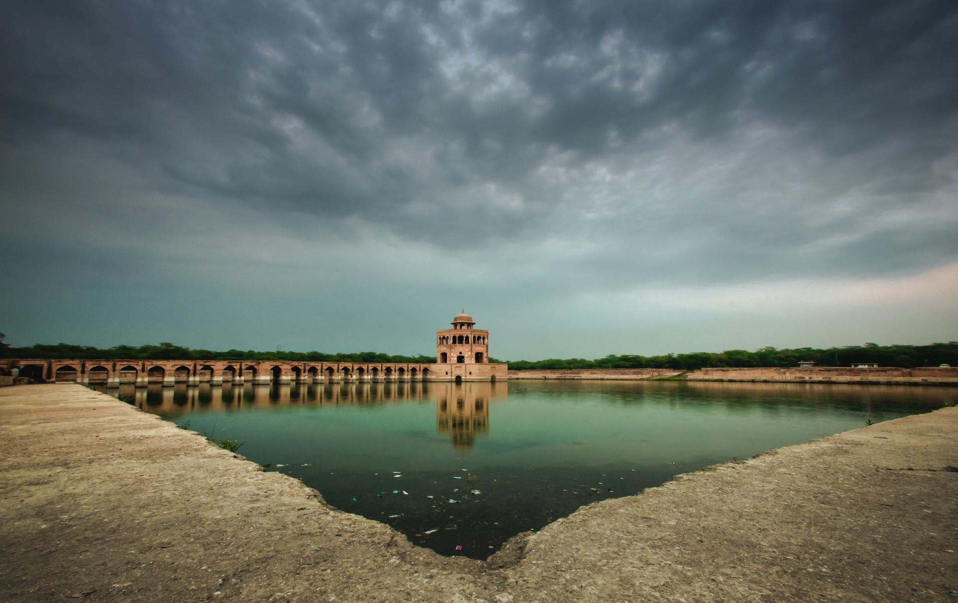 Hiran Minar in Sheikhupura, Pakistan, with dramatic cloudy skies reflecting in the calm lake.