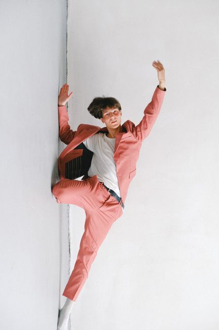 A man in a vibrant pink suit strikes a dynamic pose against a minimalist white wall.