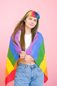 一位染着彩虹发色、手持骄傲旗帜的年轻女子在粉红色背景前微笑