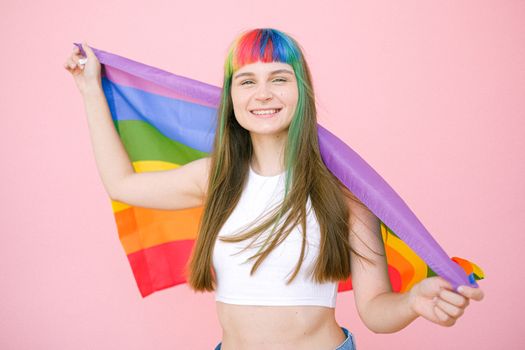 Smiling young woman with rainbow hair holding a pride flag against a pink backdrop, celebrating LGBTQ pride.