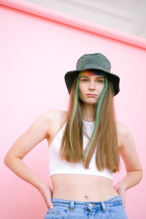 Free Portrait of a confident young woman with green hair stripes and a hat against a pink background. Stock Photo