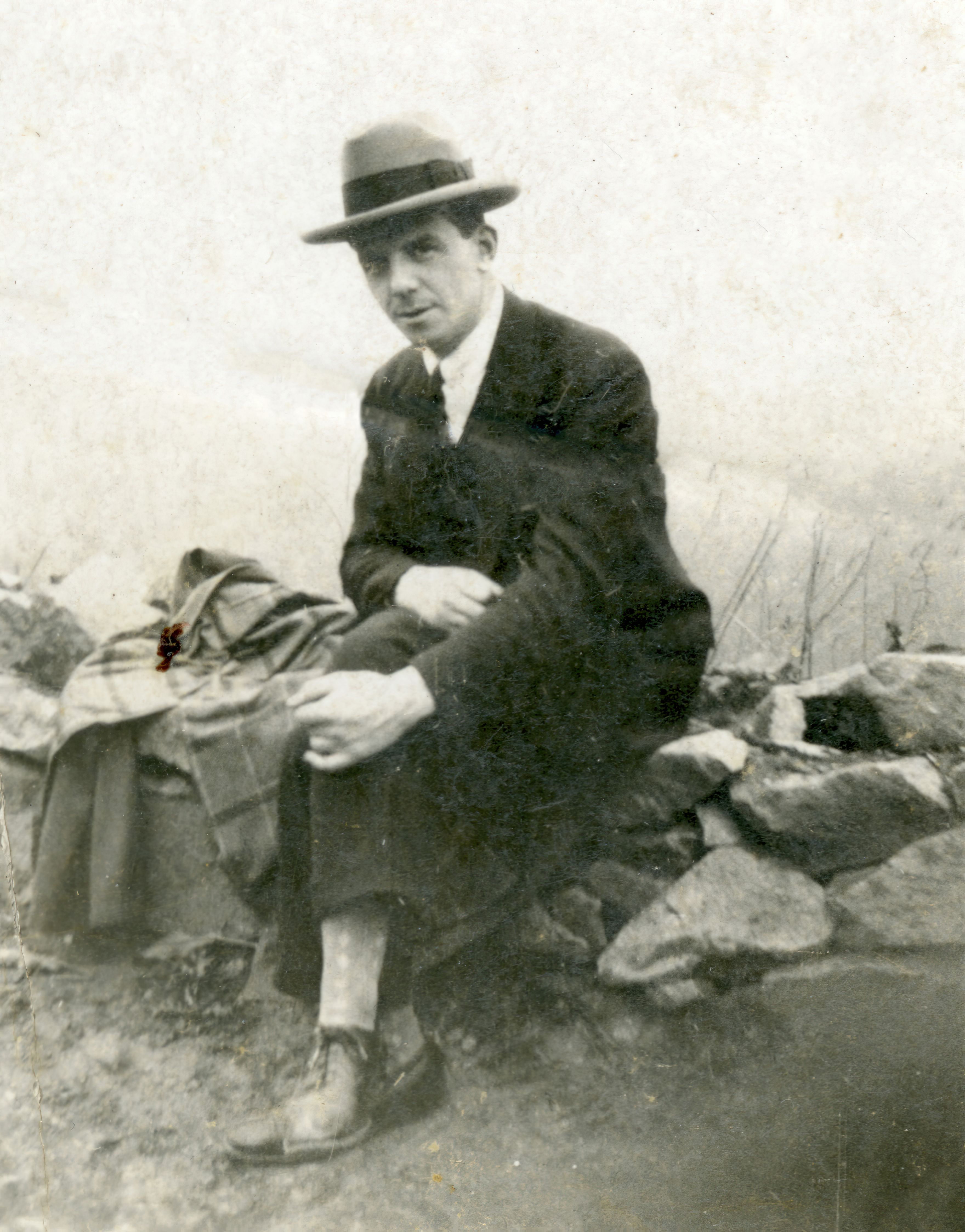 Black and white portrait of a man in a suit and hat sitting outdoors on a stone wall.