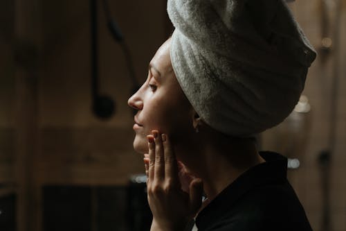 Free Woman with towel headwrap enjoying a skincare routine in dim lighting for self-care. Stock Photo
